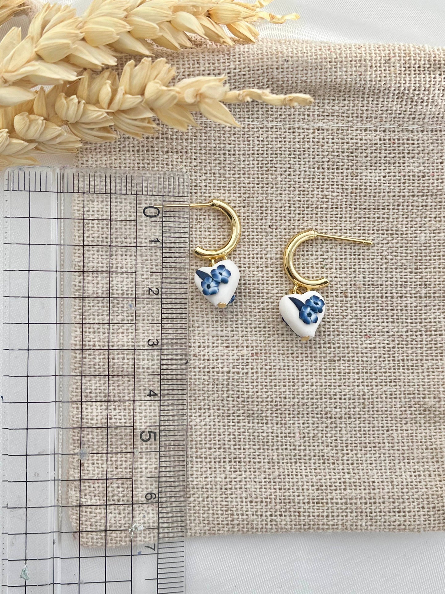 Heart shaped clay earrings with blue and white florals on a beige jewellery pouch next to a ruler