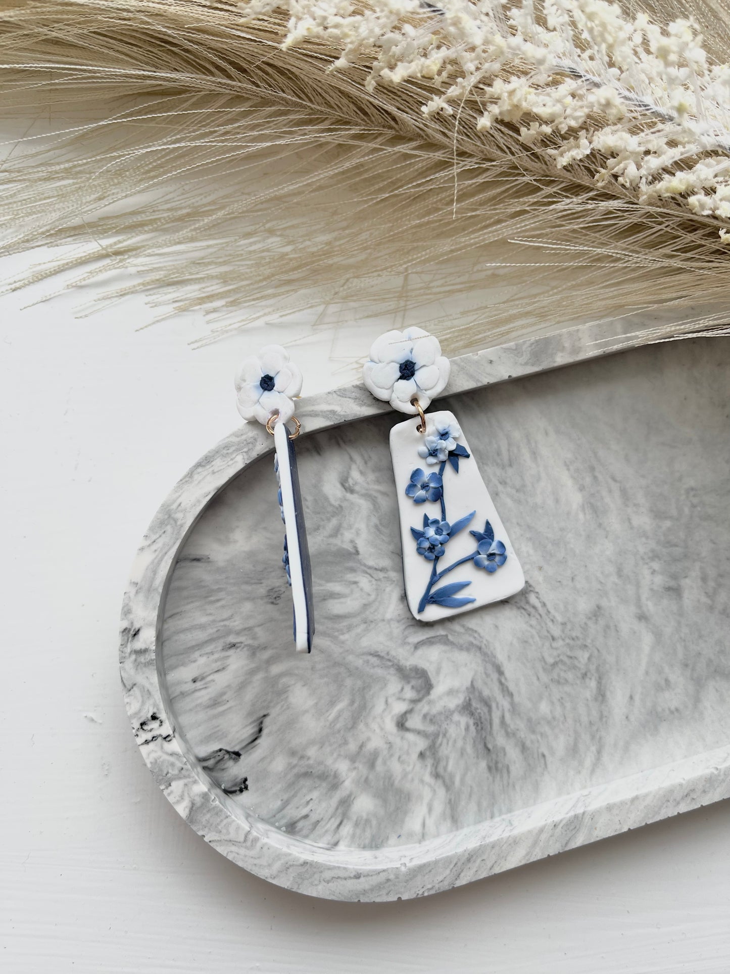 Long blue and white floral blue china clay earrings on a grey trinket dish, with one earring flipped on a side angle