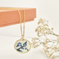 Gold clay necklace with blue and white floral pendant hanging from a jewellery box with dried flowers in the background