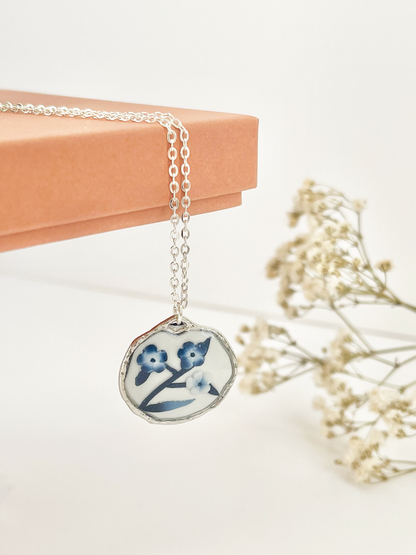 Silver clay necklace with blue and white floral pendant hanging from a jewellery box with dried flowers in the background
