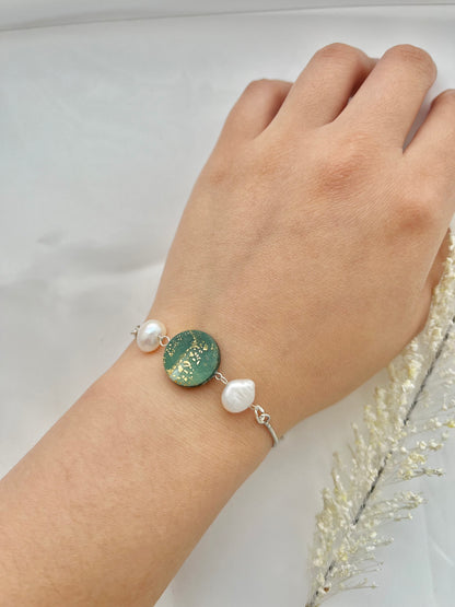 A rhodium plated bracelet with a dark green clay circle with freshwater pearls on either side on a hand