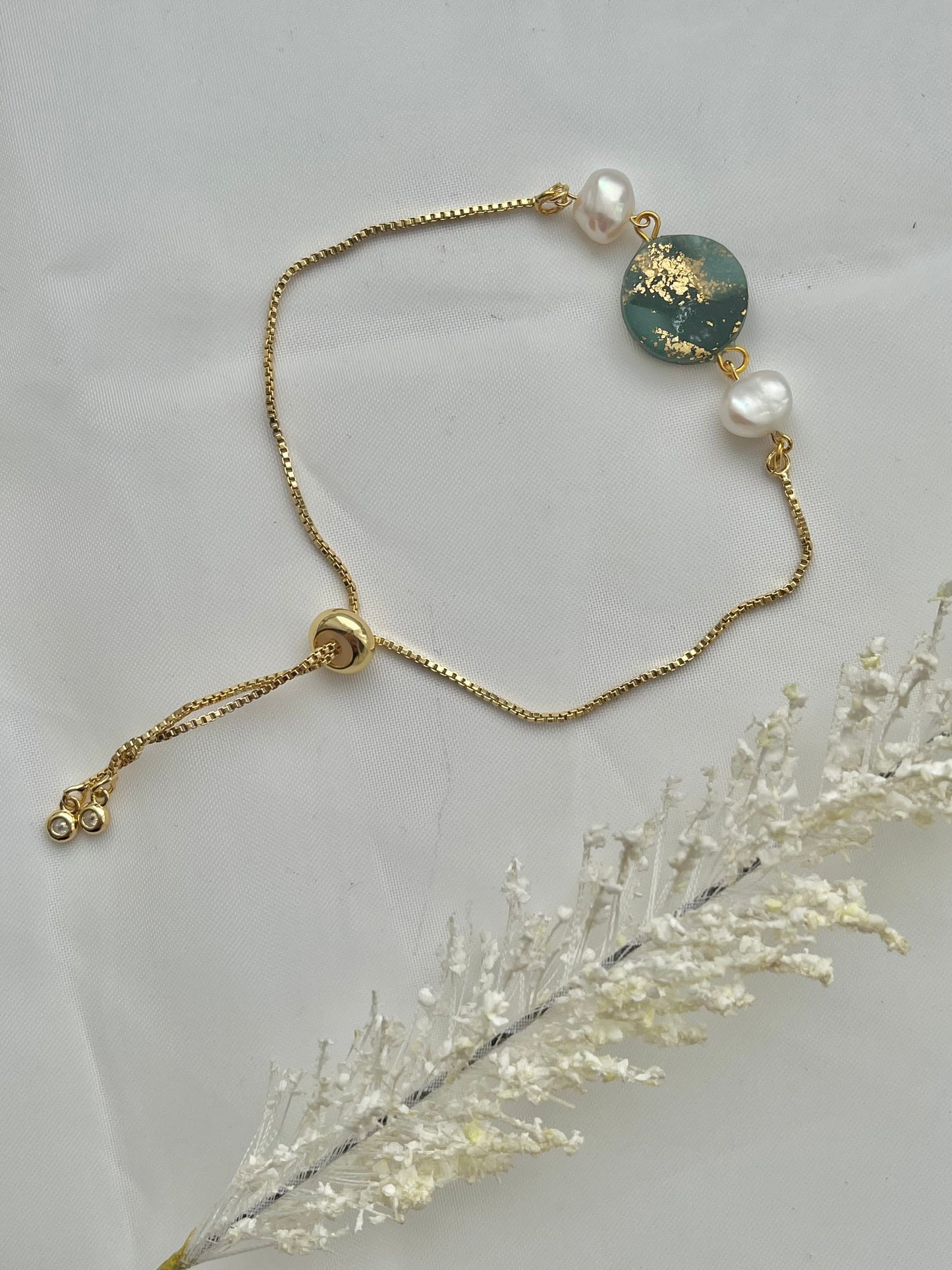 A bracelet with a dark green clay circle with freshwater pearls on either side laid flat down to show the adjustable slider