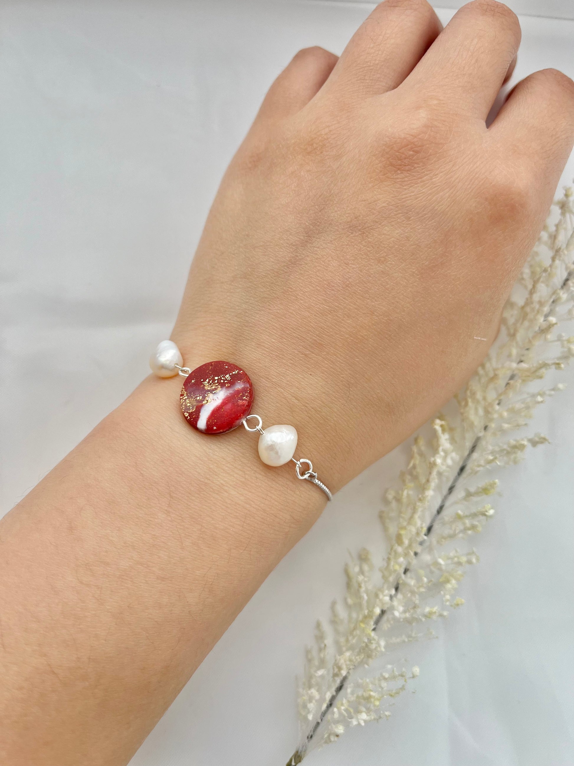 A rhodium plated bracelet with a dark red clay circle with freshwater pearls on either side on a hand