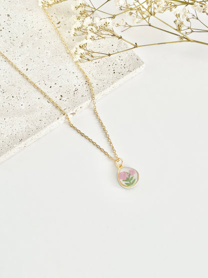 A gold clay necklace with a small carnation pendant laid on a flat white surface next to dried flowers
