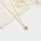 A gold clay necklace with a small pendant with violets laid on a white surface next to dried flowers