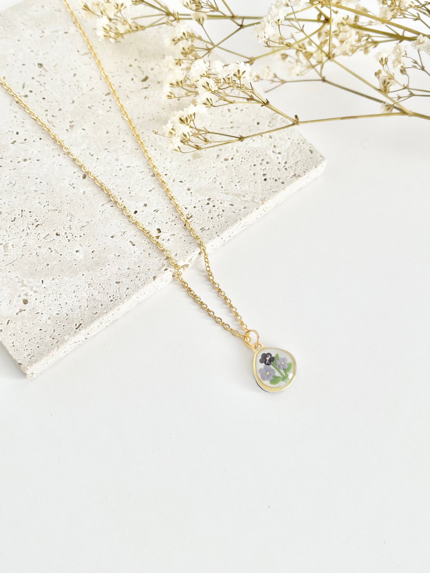 A gold clay necklace with a small pendant with violets laid on a white surface next to dried flowers
