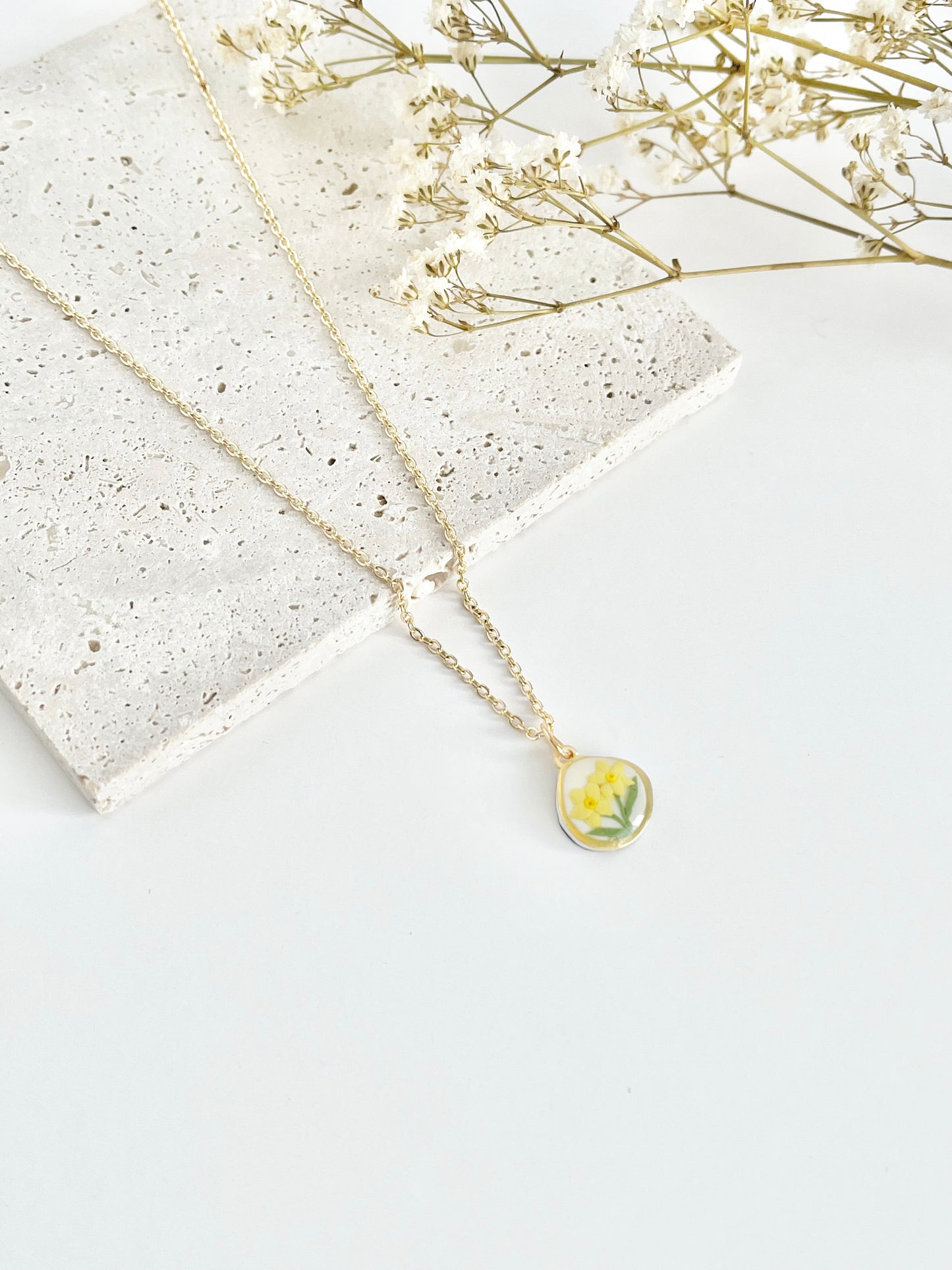 Gold clay necklace with a small daffodil pendant on a white surface with dried flowers.