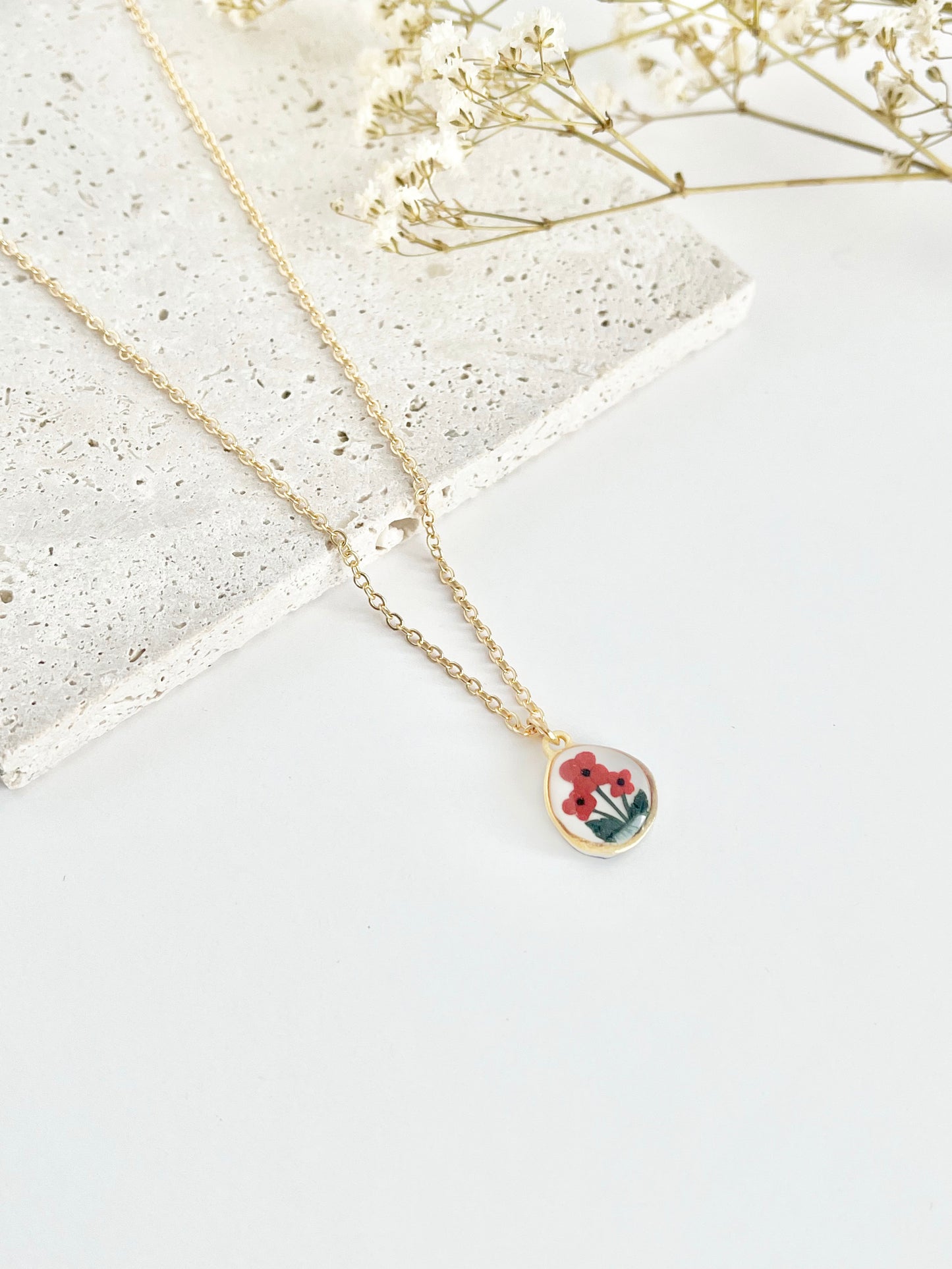 Gold clay necklace with a small pendant featuring poppies on a light surface next to dried flowers