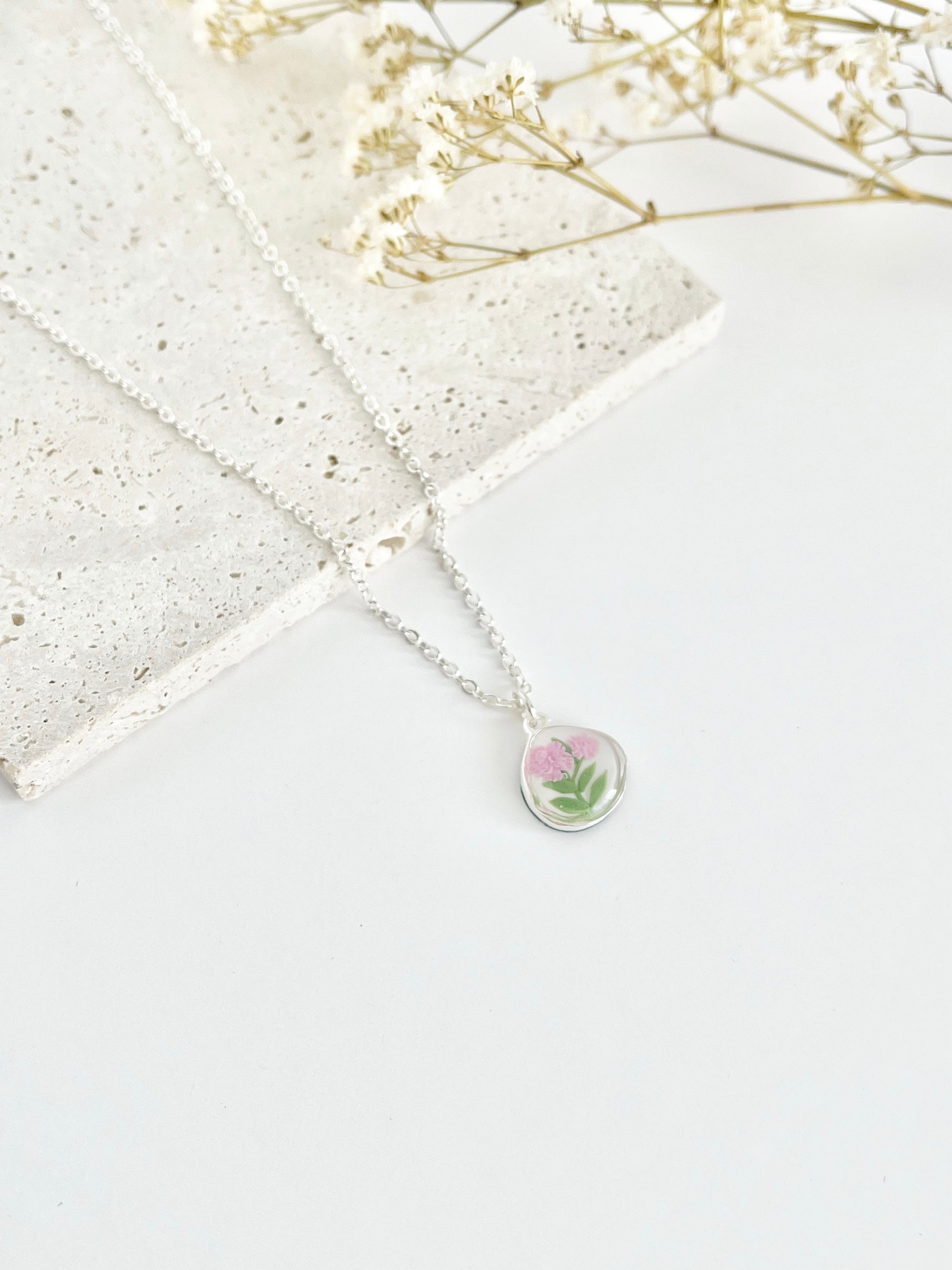 A silver clay necklace with a small carnation pendant laid on a flat white surface next to dried flowers