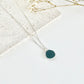 A silver clay necklace with a small green pendant laid on a flat white surface next to dried flowers