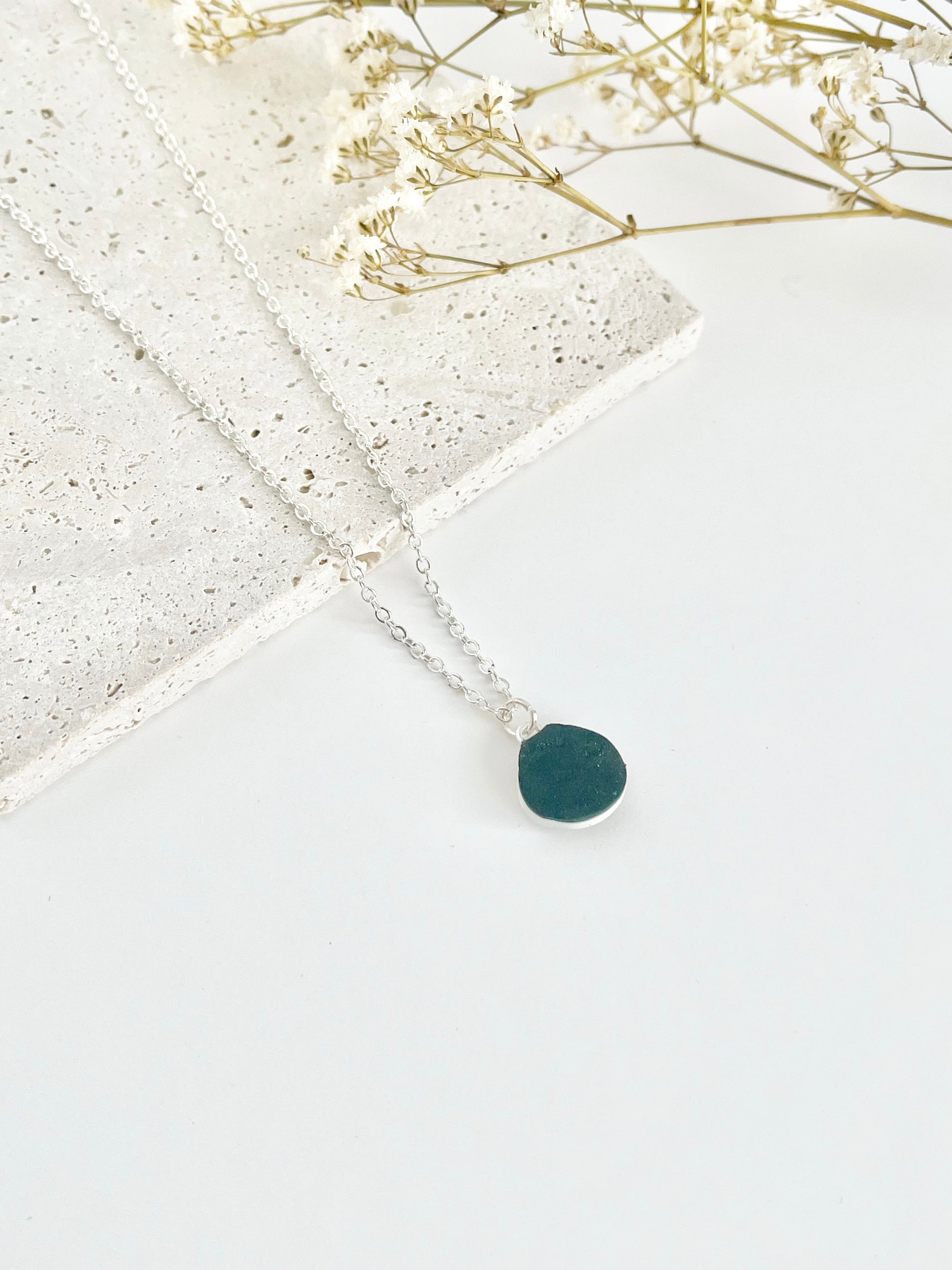 A silver clay necklace with a small green pendant laid on a flat white surface next to dried flowers