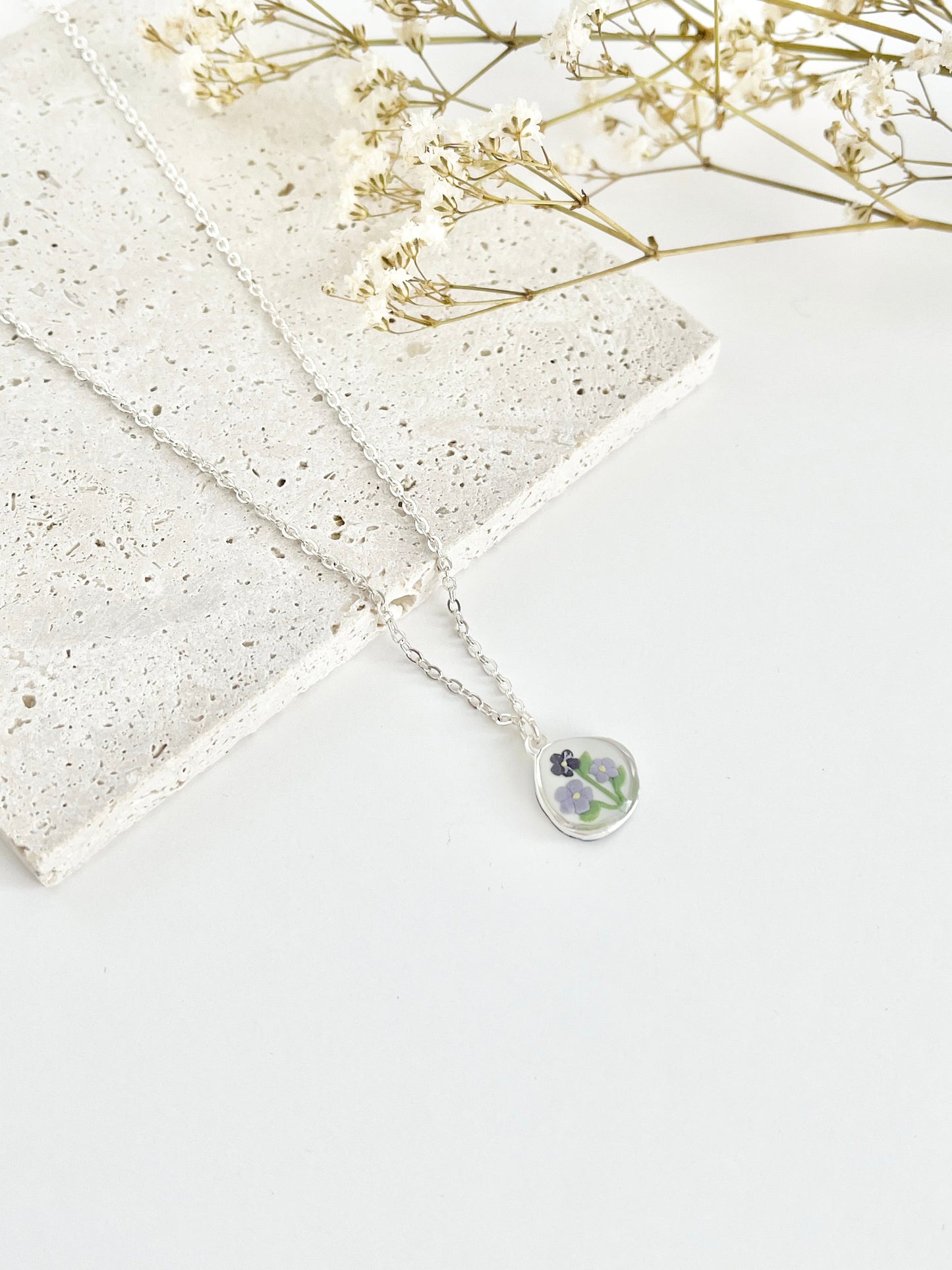 A silver clay necklace with a small pendant with violets laid on a white surface next to dried flowers