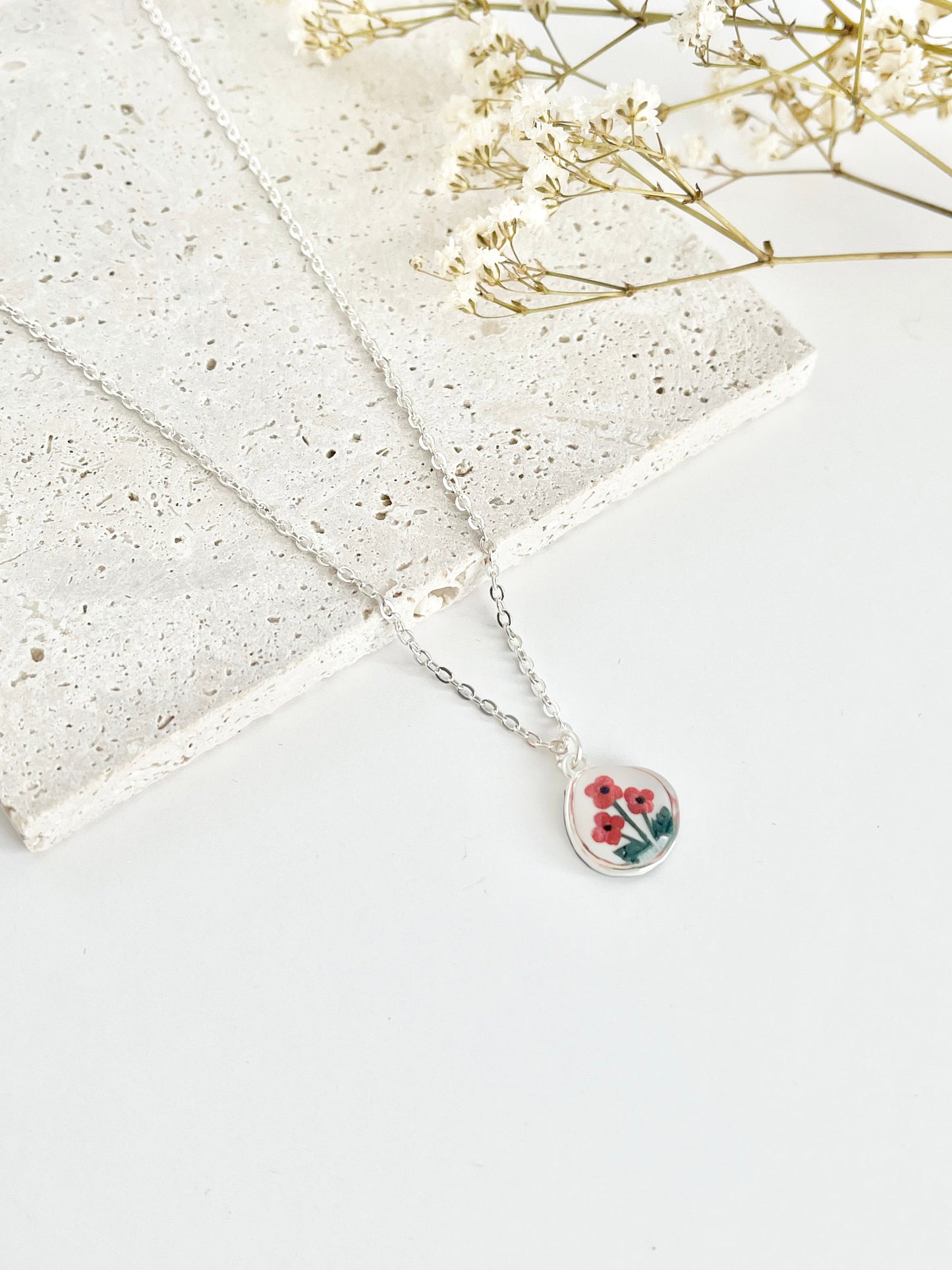 Silver clay necklace with a small pendant featuring poppies on a light surface next to dried flowers