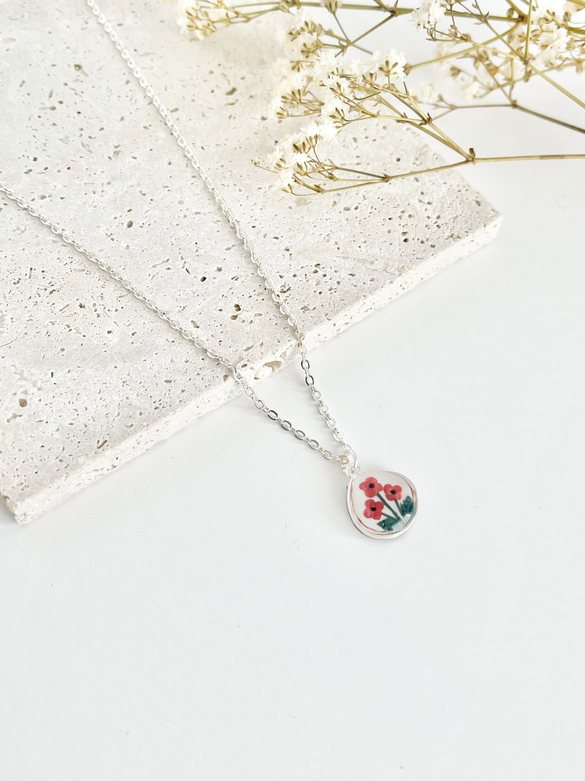 Silver clay necklace with a small pendant featuring poppies on a light surface next to dried flowers