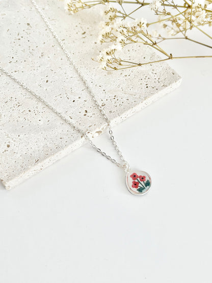 Silver clay necklace with a small pendant featuring poppies on a light surface next to dried flowers