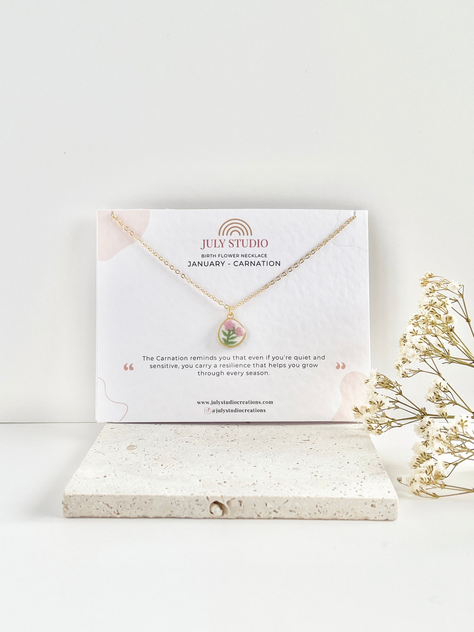 A gold clay necklace with a small carnation pendant on a jewellery card against a white background next to dried flowers