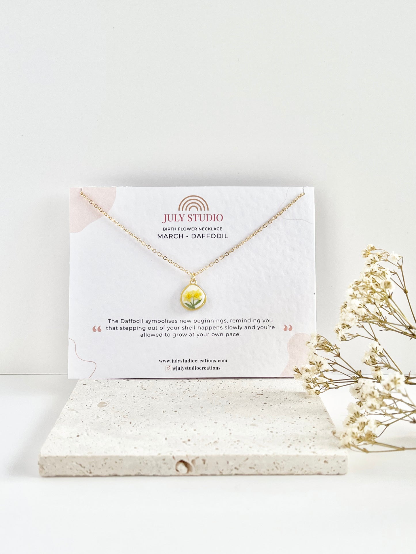 Gold clay necklace with a small daffodil pendant on a jewellery card, placed on a white surface with a light background, next to dried flowers.