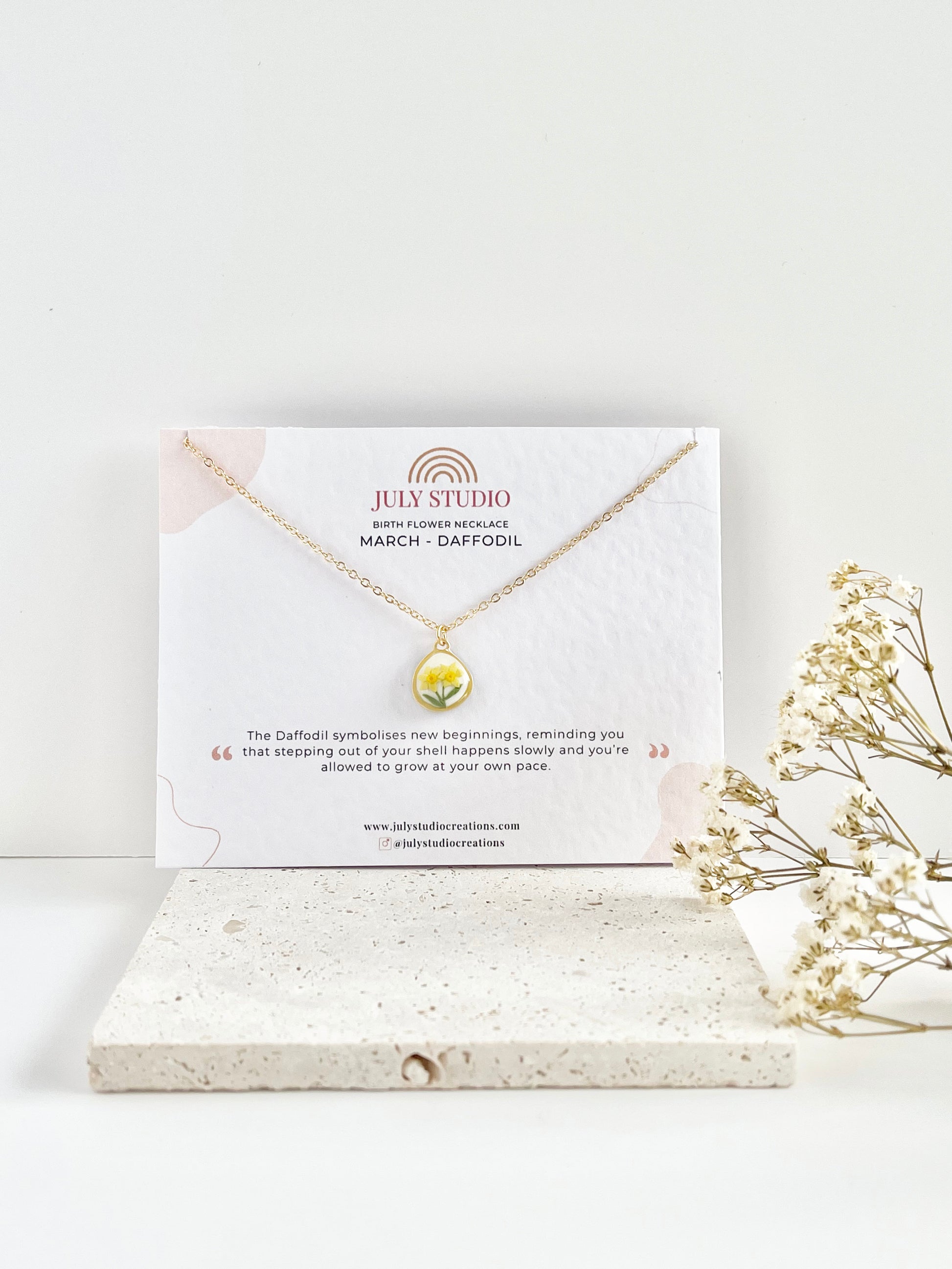 Gold clay necklace with a small daffodil pendant on a jewellery card, placed on a white surface with a light background, next to dried flowers.
