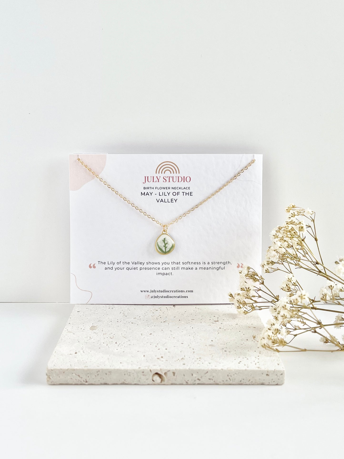 Gold clay necklace with a lily of the valley pendant on a jewellery card on top of a textured tile, with dried flowers beside it.