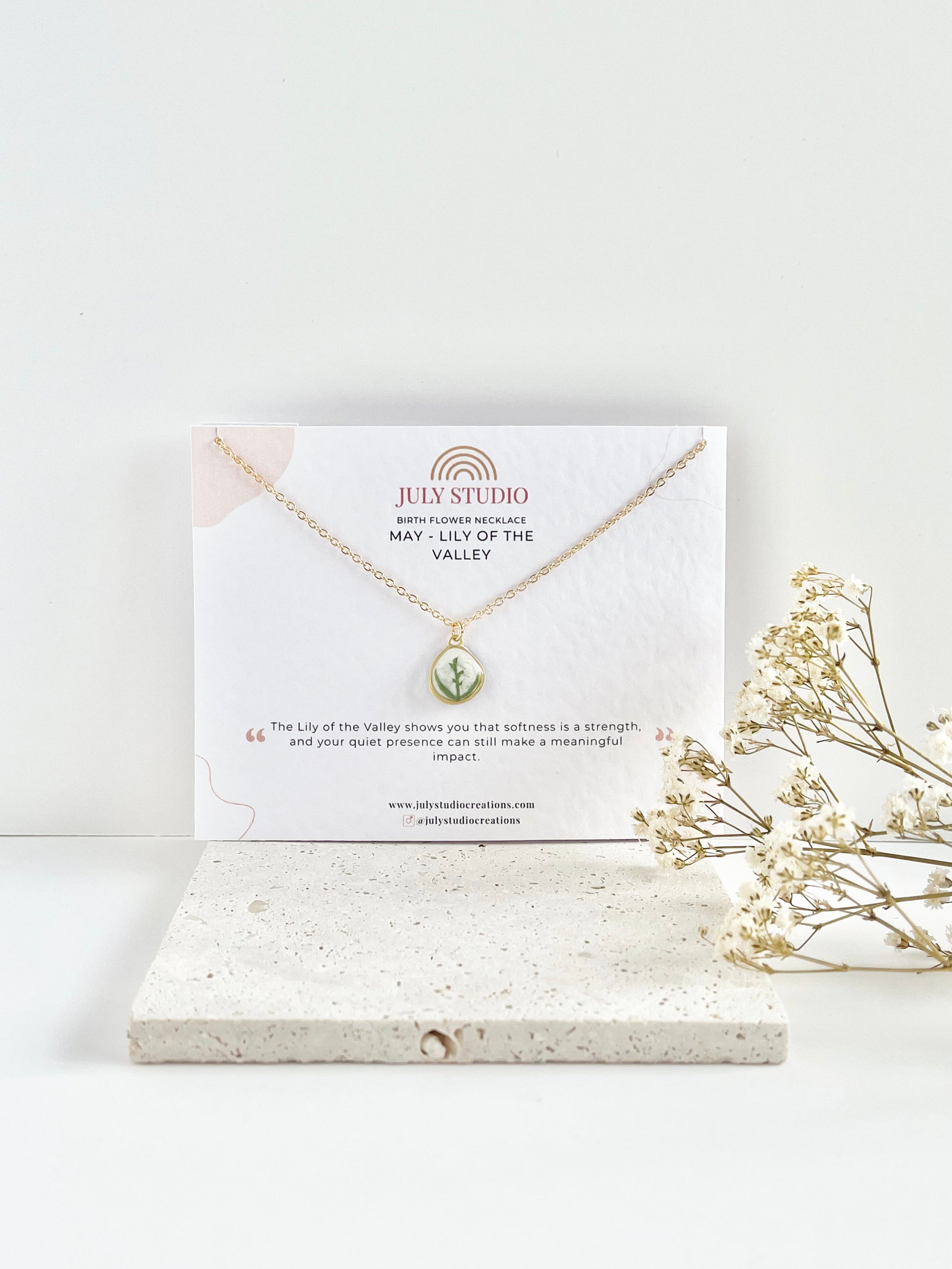 Gold clay necklace with a lily of the valley pendant on a jewellery card on top of a textured tile, with dried flowers beside it.