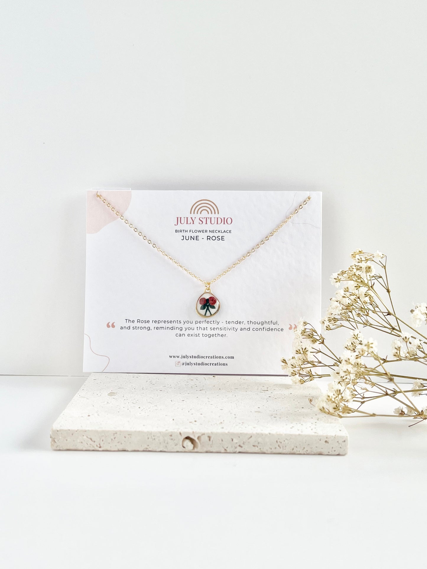 Gold clay necklace with a rose pendant on a jewellery card, placed on a textured tile with dried flowers.