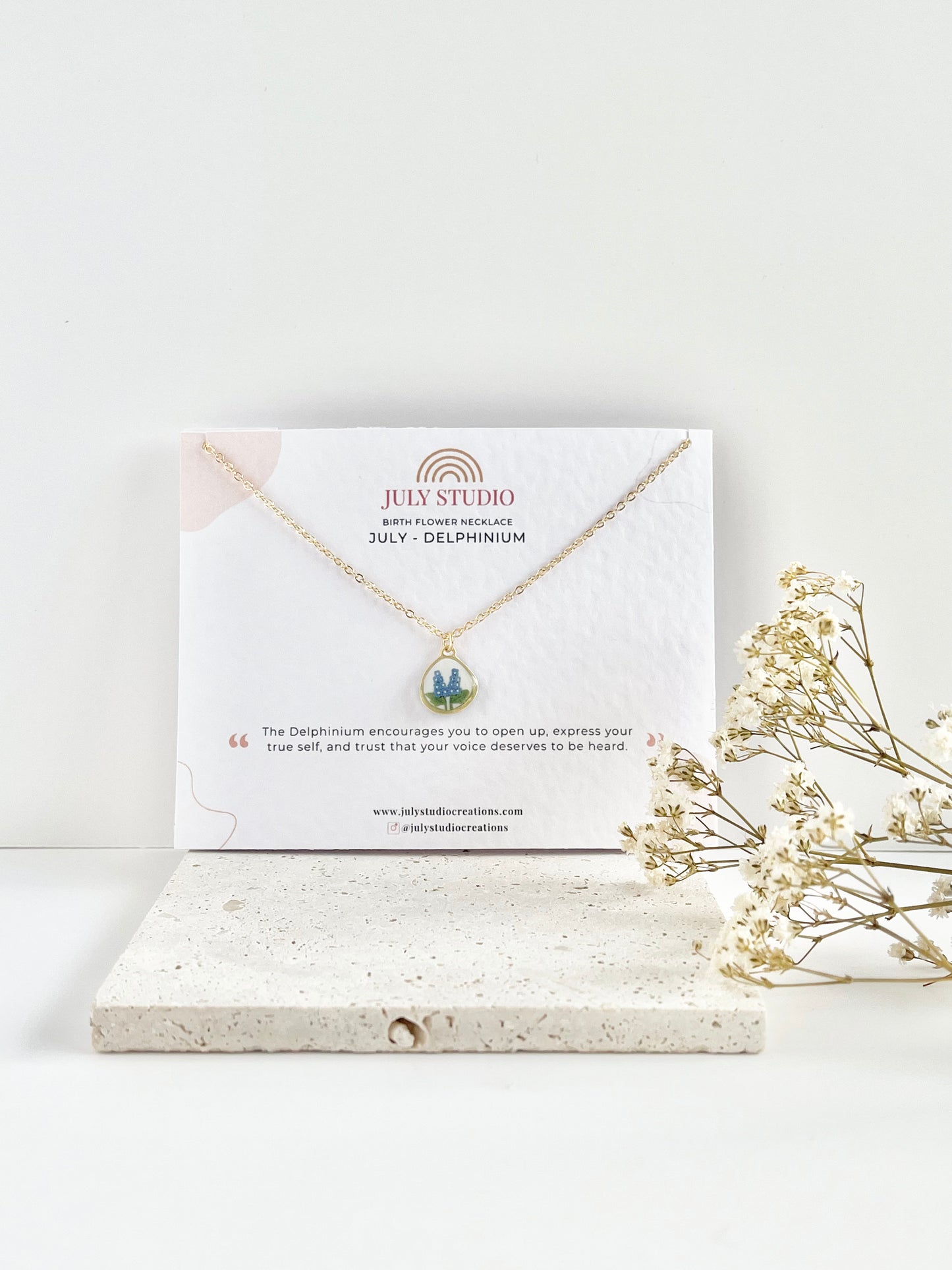 Gold clay necklace with floral pendant on a jewellery card, placed on a white surface with dried flowers.