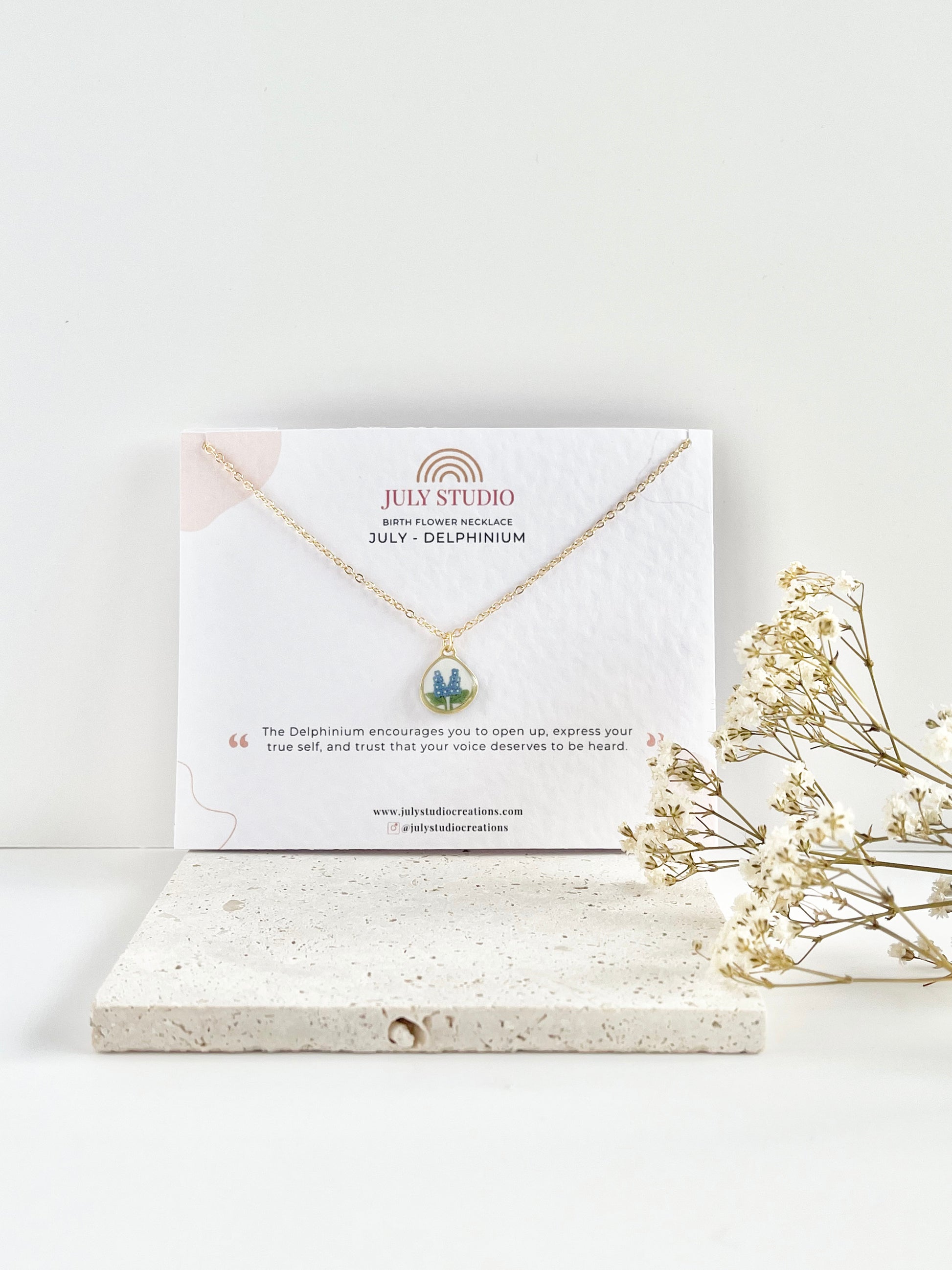 Gold clay necklace with floral pendant on a jewellery card, placed on a white surface with dried flowers.