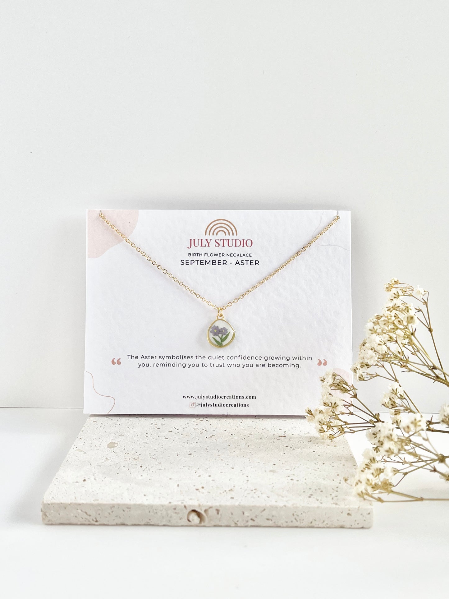Gold clay necklace with a floral pendant on a white card with 'July Studio' branding, placed on a light surface with dried flowers.