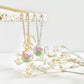 A gold and a silver clay necklace hanging from a stone tile with small pendants with pink carnations and dried flowers in the background