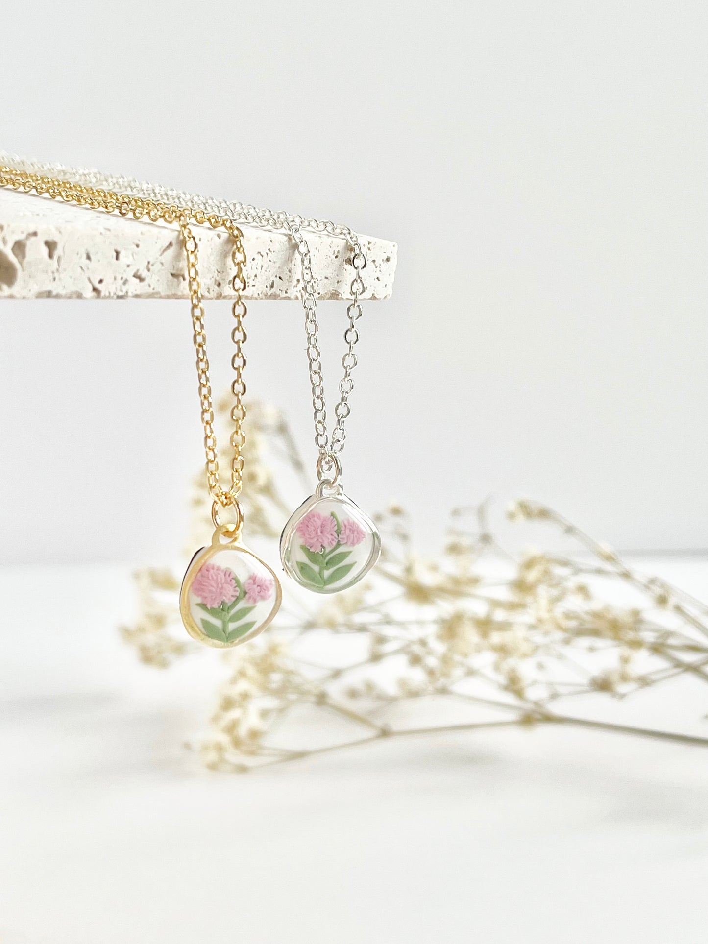 A gold and a silver clay necklace hanging from a stone tile with small pendants with pink carnations and dried flowers in the background