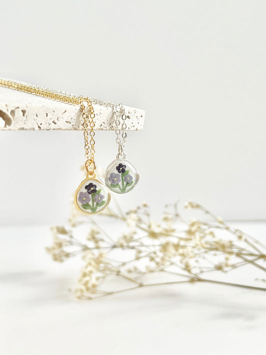 A gold and a silver necklace with a small pendant with violets hanging from a beige tile with dried flowers in the background