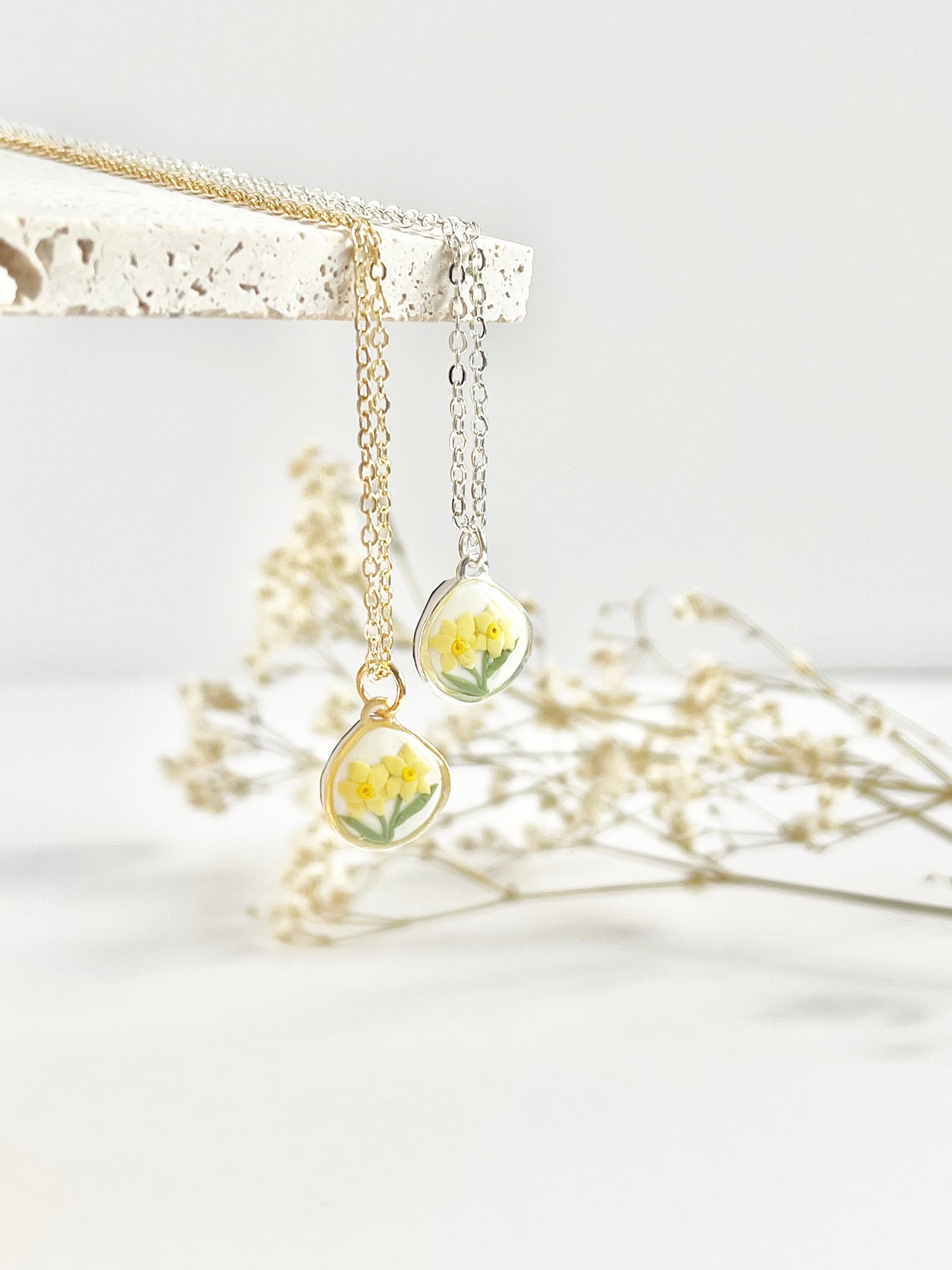 Gold necklace and silver clay necklace with daffodil pendant hanging from a tile against a white background with dried flowers in the background