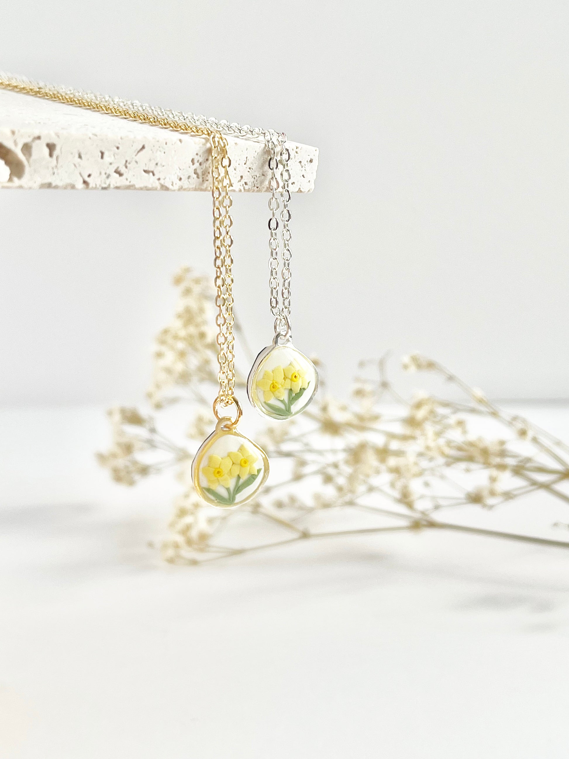 Gold necklace and silver clay necklace with daffodil pendant hanging from a tile against a white background with dried flowers in the background