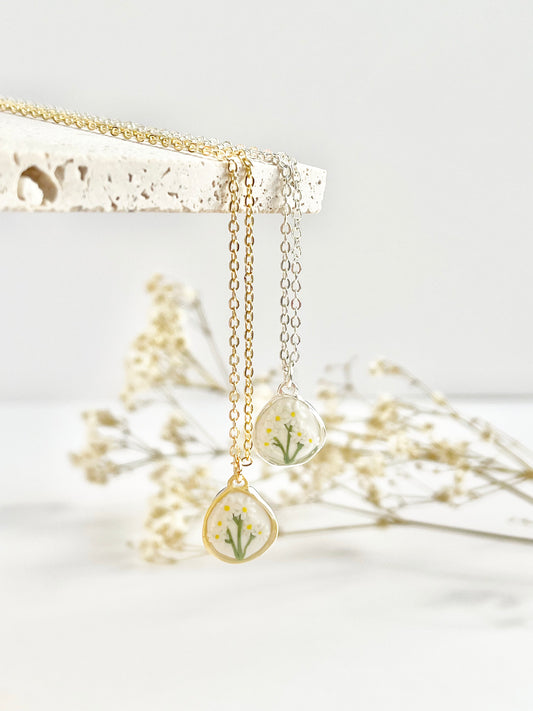 Dainty necklaces in gold and silver with a teardrop pendant with daises featuring dried flowers on a white background.
