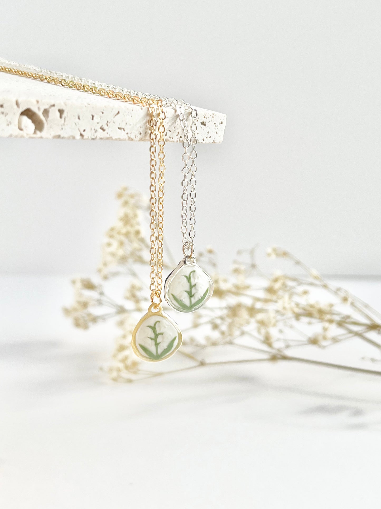 A gold and silver clay necklace with teardrop pendants hanging from a textured tile and dried flowers in the background