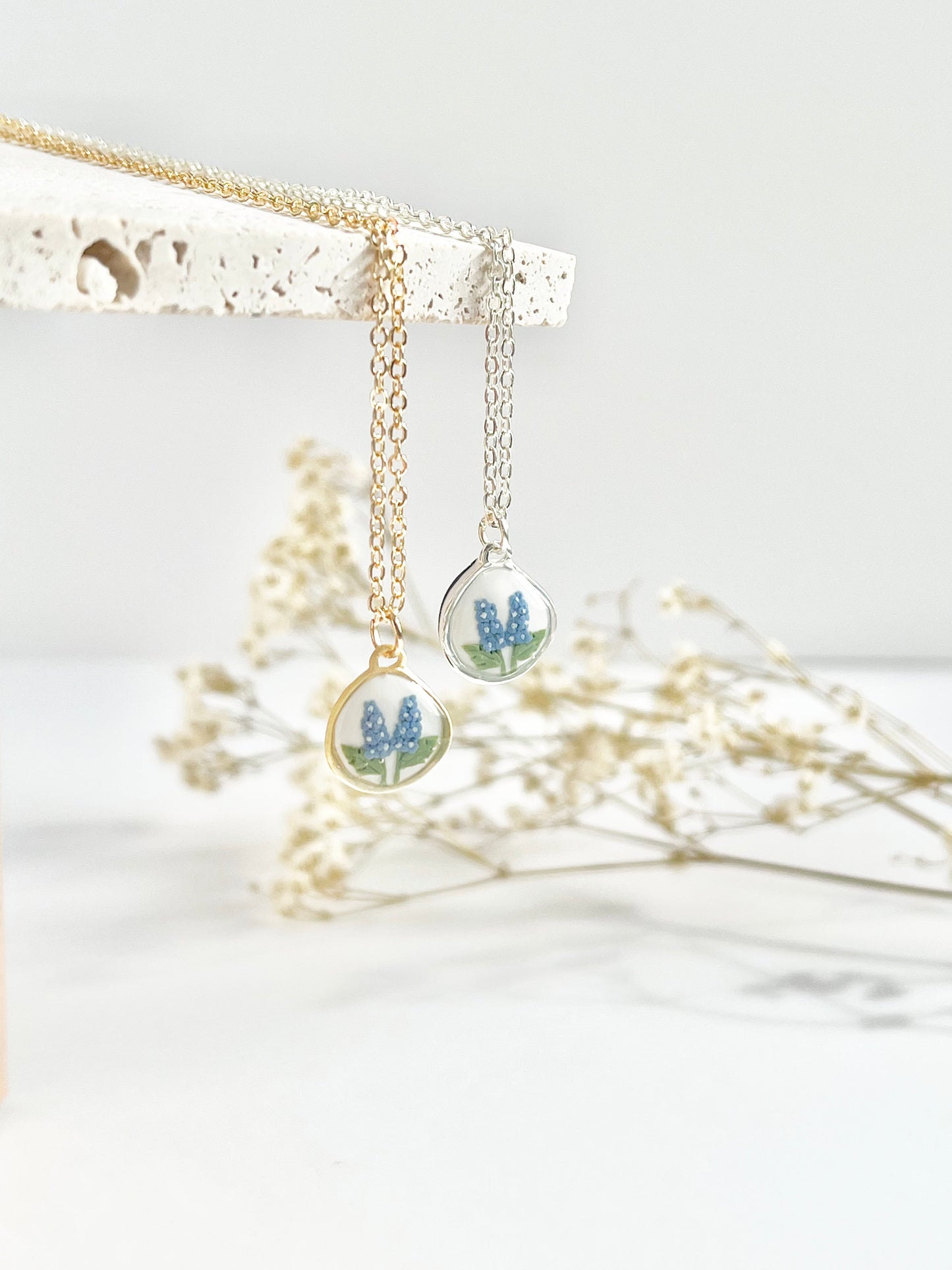 Gold necklace and silver clay necklace with a pendant featuring small flowers hanging from a textured tile against a white background with dried flowers