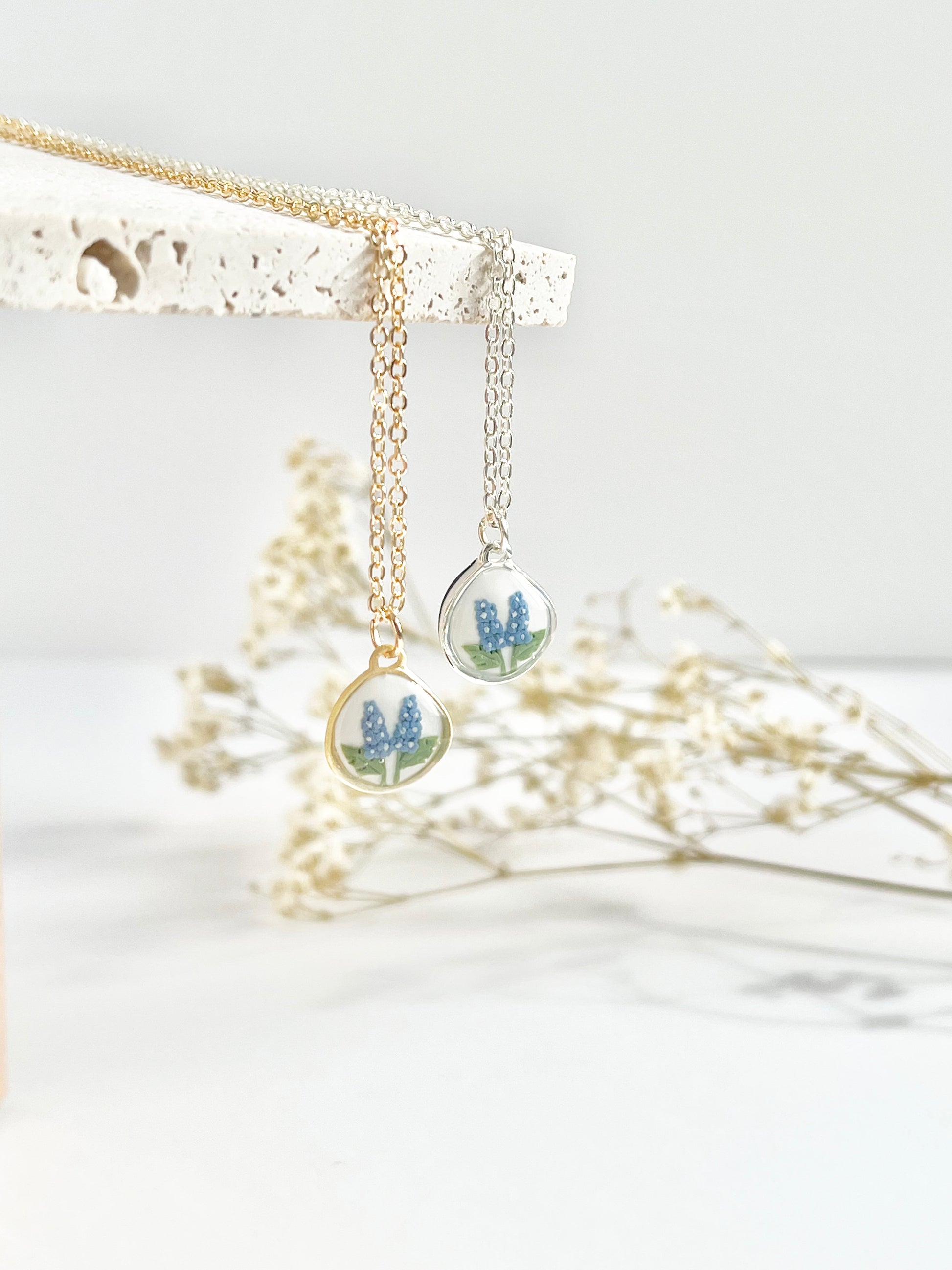 Gold necklace and silver clay necklace with a pendant featuring small flowers hanging from a textured tile against a white background with dried flowers