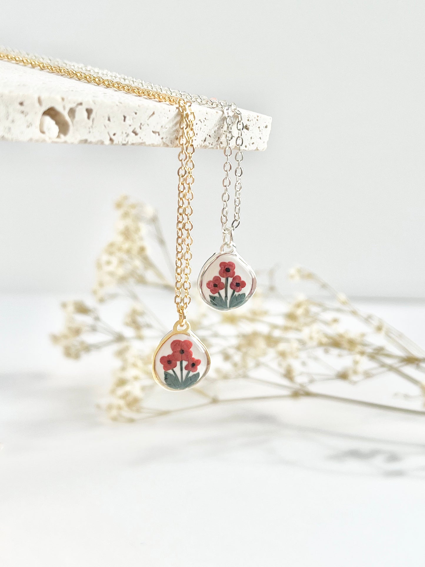 Gold clay necklace and silver clay necklace with small pendant featuring poppies hanging from a textured tile against a white background with dried flowers in the background