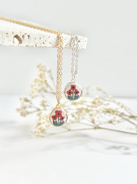 Gold necklace and silver necklace with small pendant featuring poppies hanging from a textured tile against a white background with dried flowers in the background