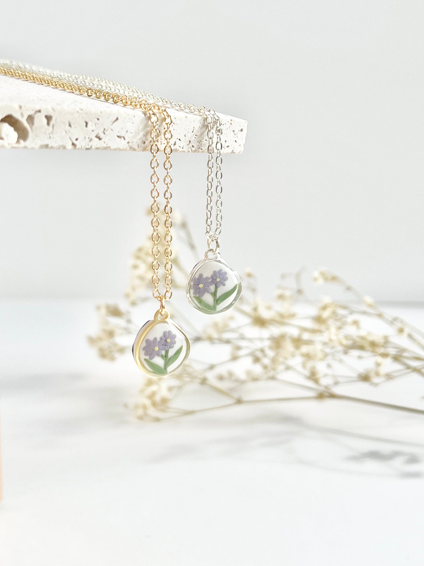Gold and silver clay necklaces with a pendant featuring purple flowers hanging from a textured tile with dried flowers in the background