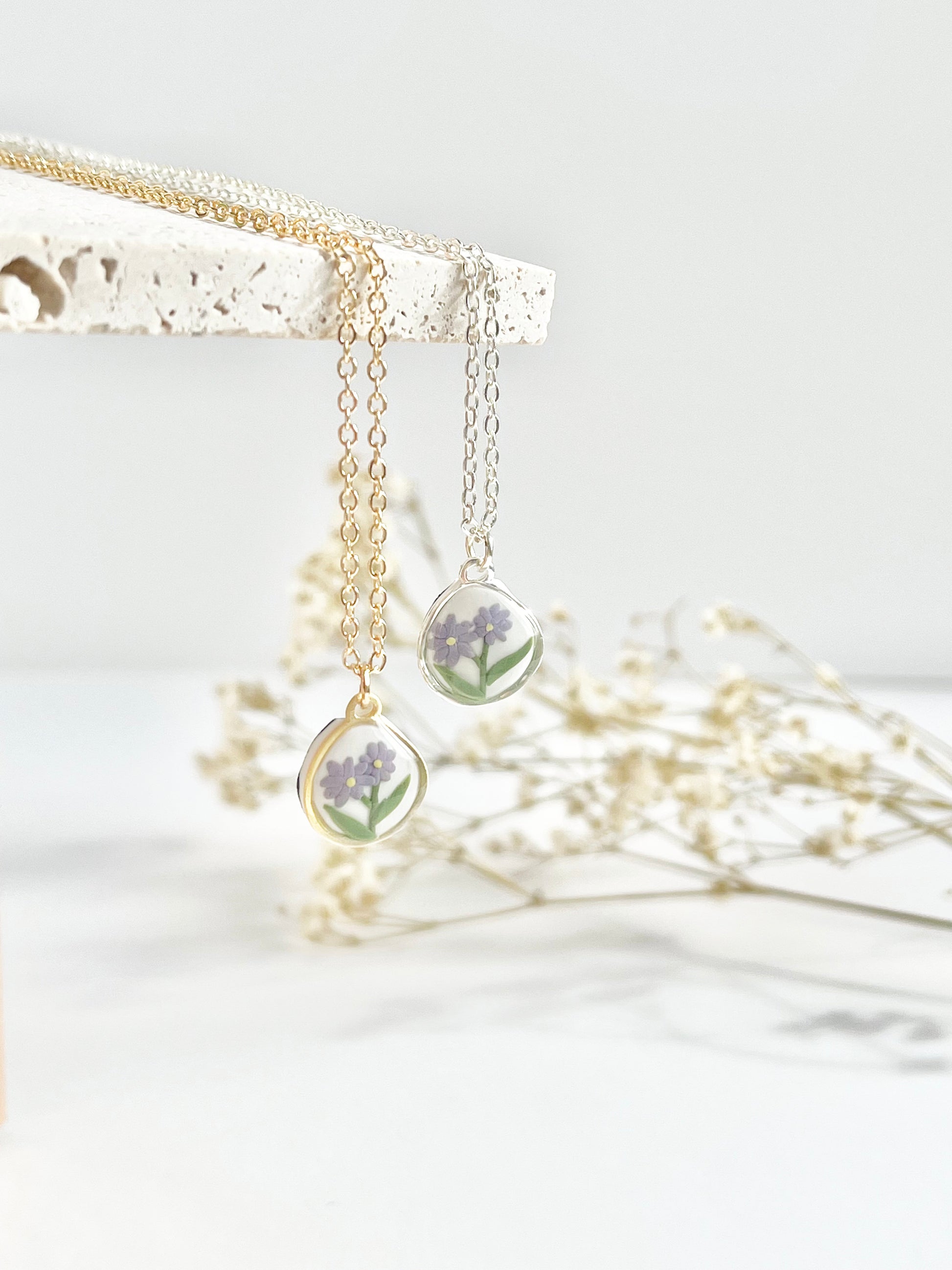 Gold and silver clay necklaces with a pendant featuring purple flowers hanging from a textured tile with dried flowers in the background