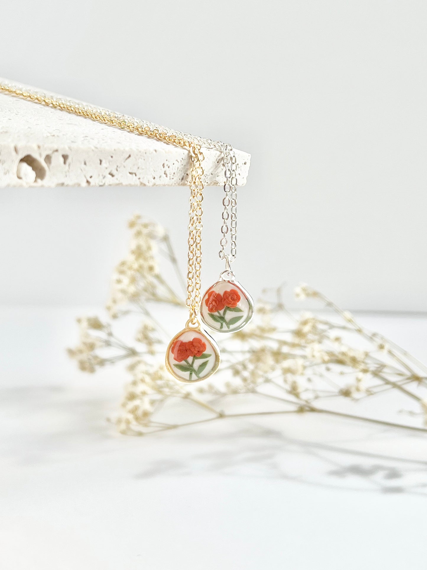 Gold and silver dainty clay necklaces hanging from a textured tile with small pendants featuring orange marigolds
