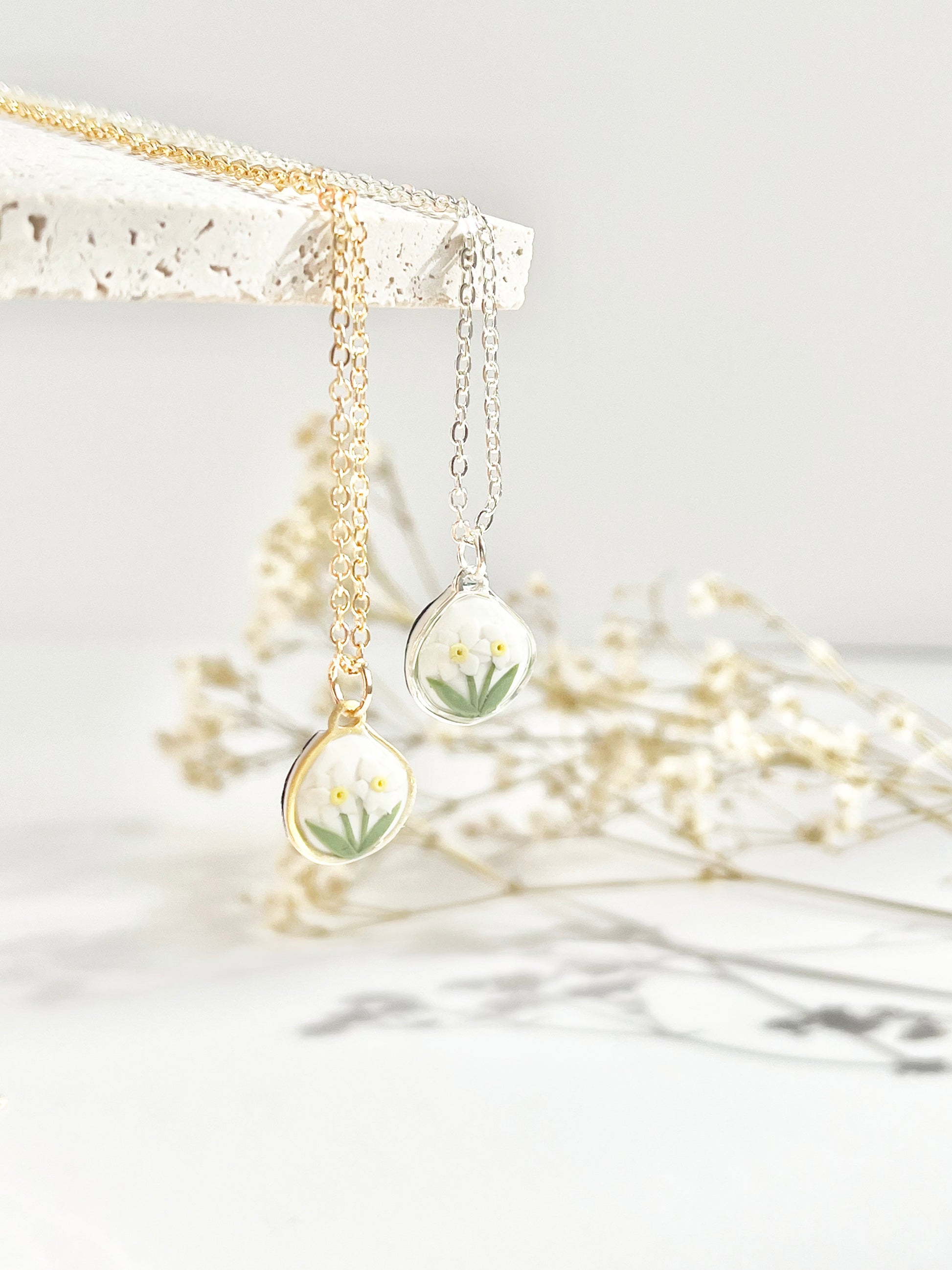 Gold and silver dainty clay necklaces with floral pendants hanging from a textured tile against a white background