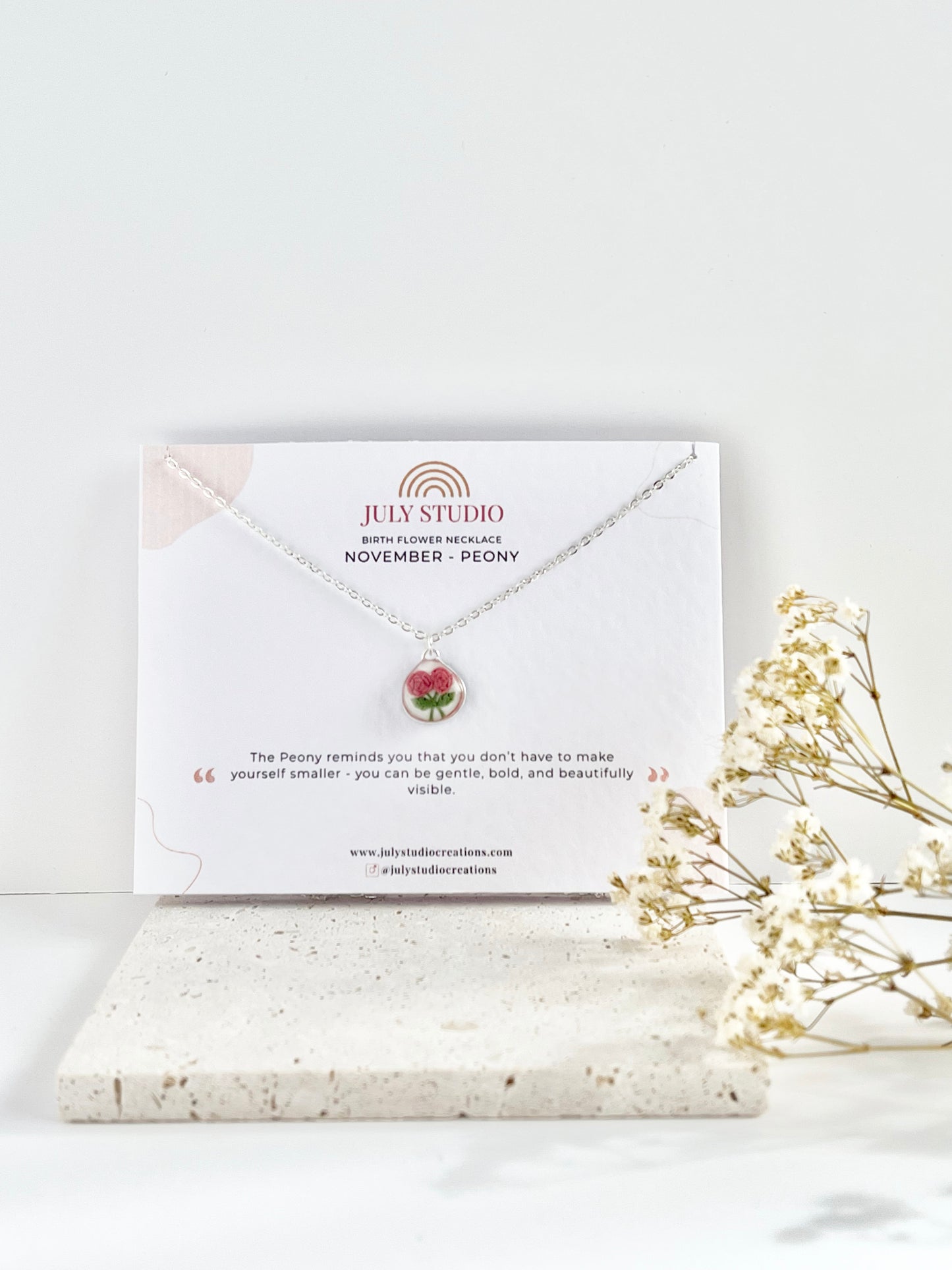 Silver clay necklace with a floral pendant on a branded card, next to dried flowers on a light background