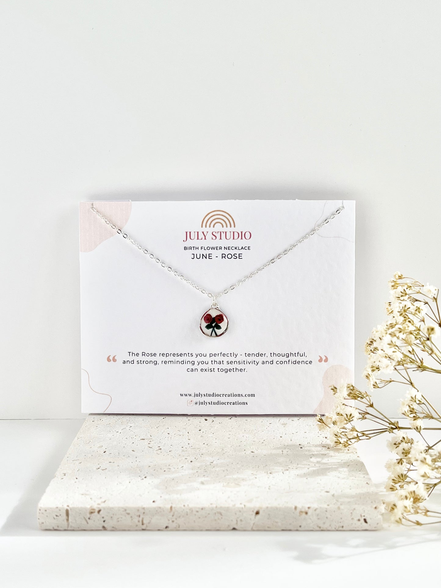 Silver clay necklace with a rose pendant on a jewellery card, placed on a textured tile with dried flowers.