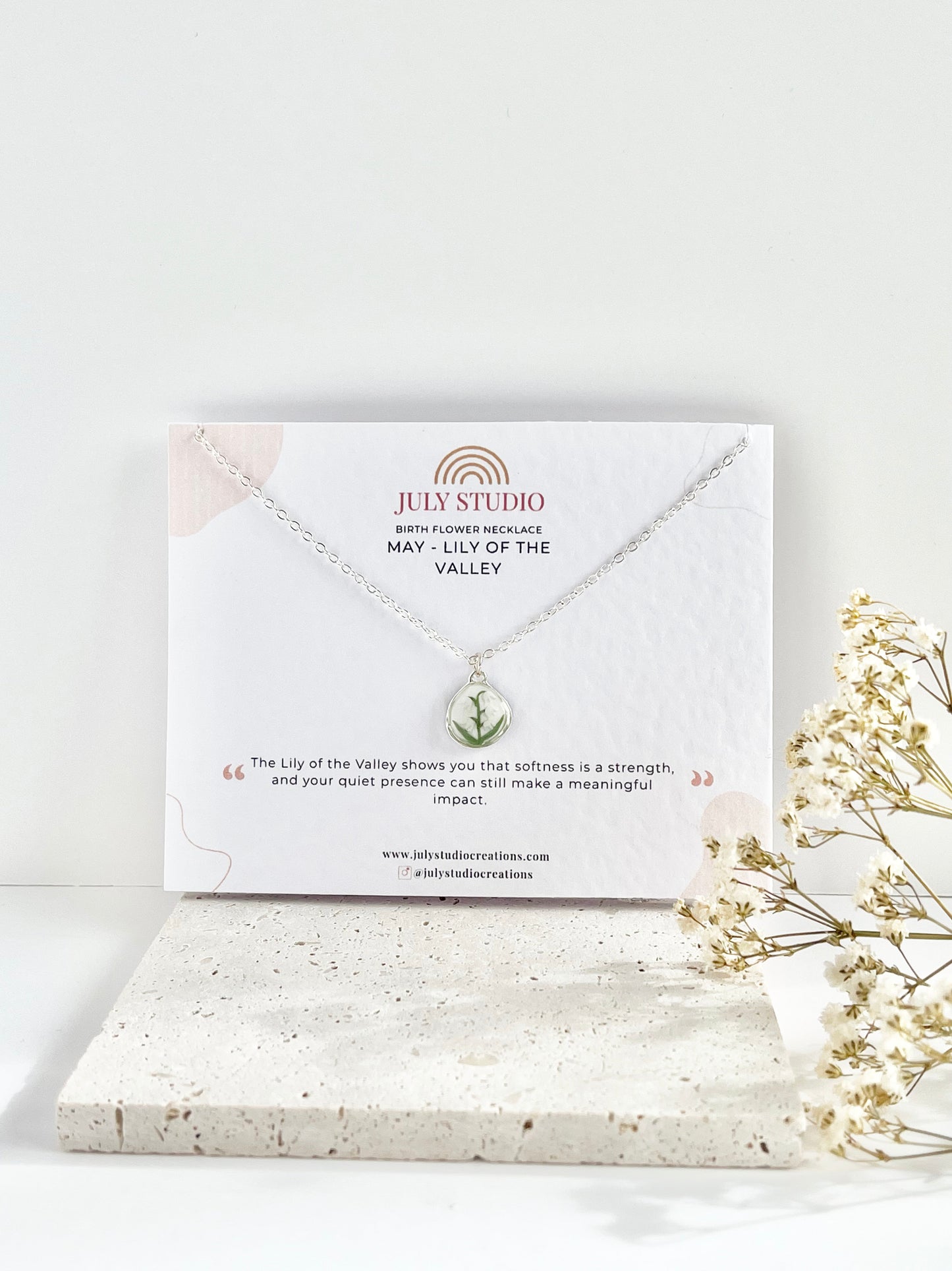 Silver clay necklace with a lily of the valley pendant on a jewellery card on top of a textured tile, with dried flowers beside it.
