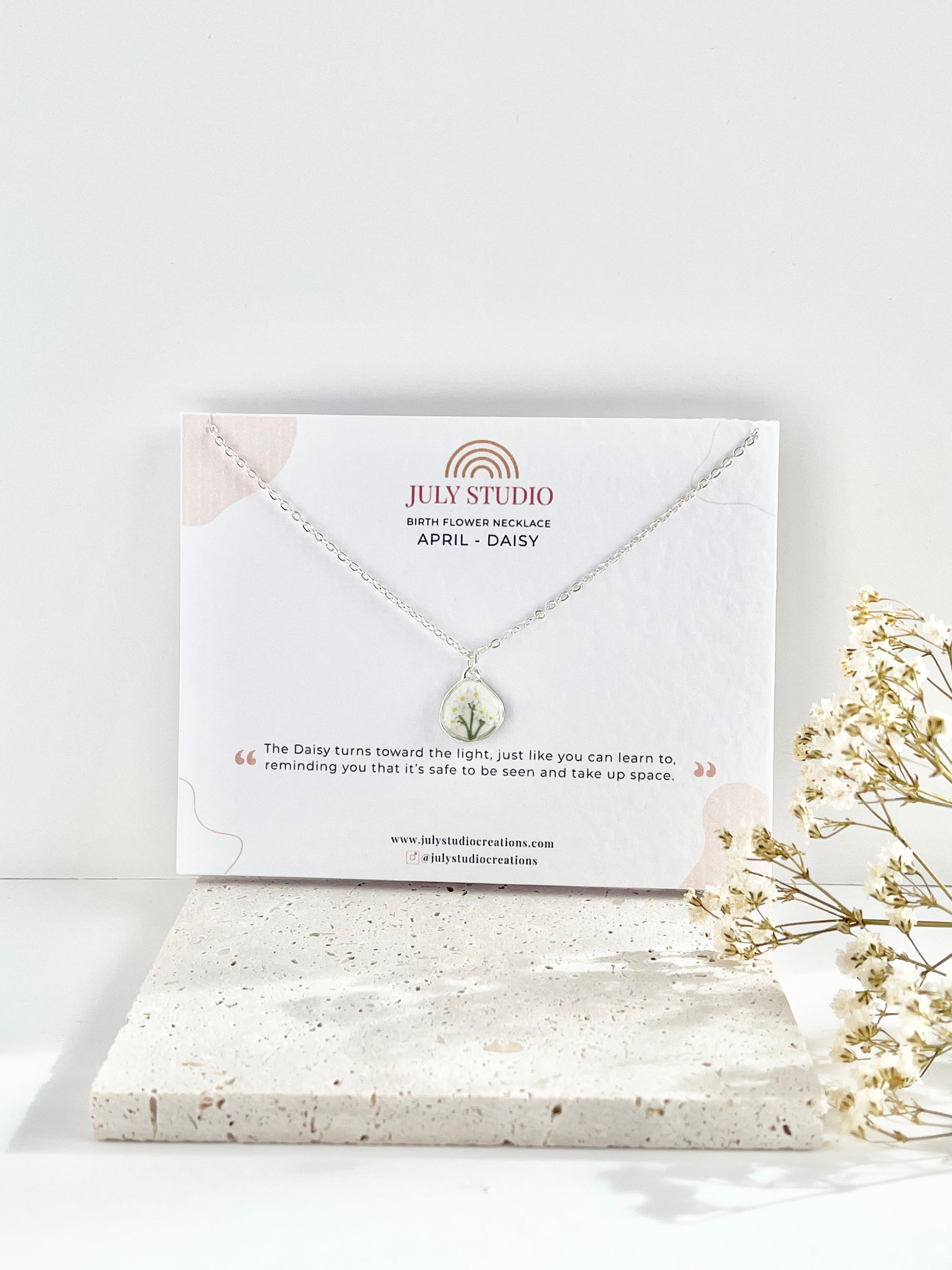 Silver necklace with a daisy pendant on a July Studio jewellery card, next to dried flowers on a light background