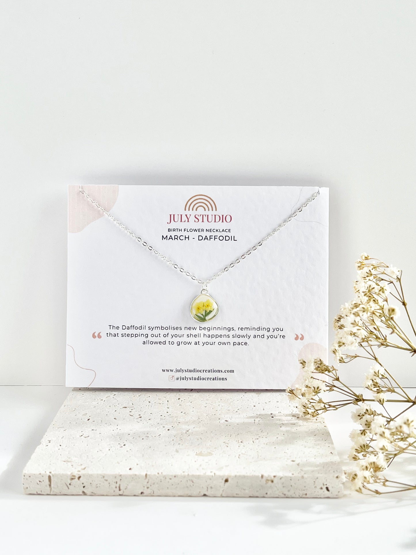 Silver clay necklace with a small daffodil pendant on a jewellery card, placed on a white surface with a light background, next to dried flowers.