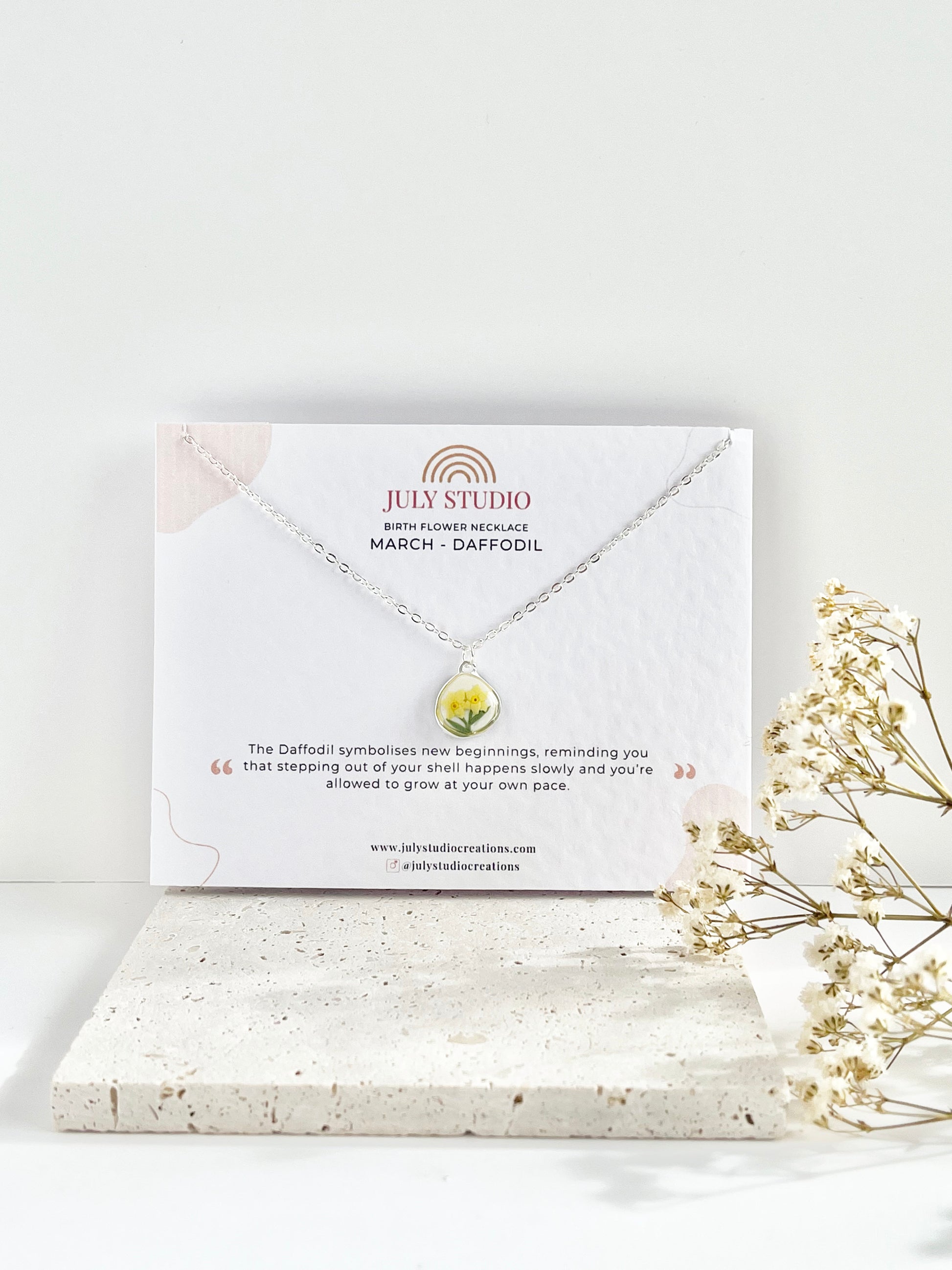 Silver clay necklace with a small daffodil pendant on a jewellery card, placed on a white surface with a light background, next to dried flowers.