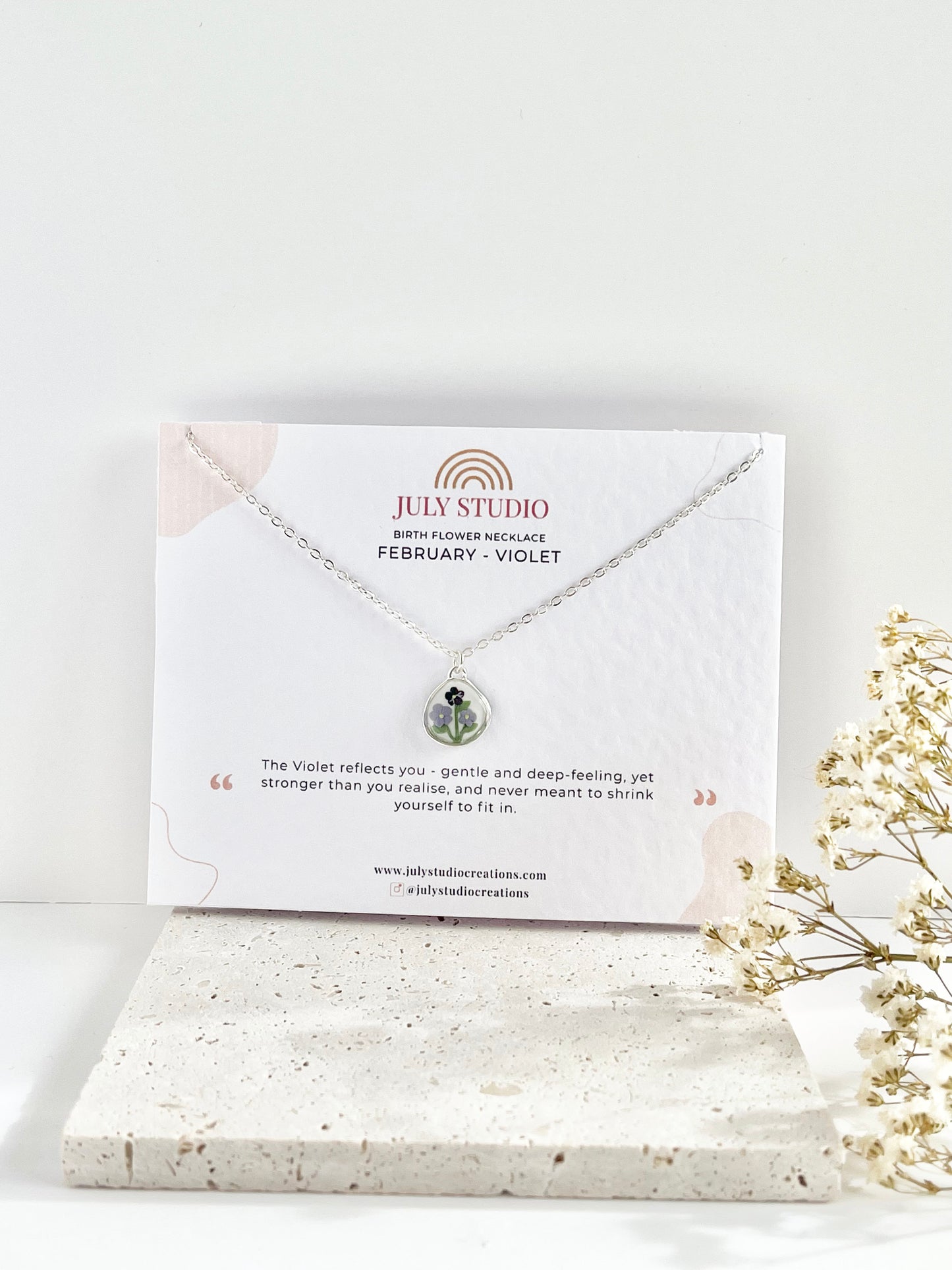 A silverc lay necklace with a small pendant with violets on a jewellery card against a white background next to dried flowers